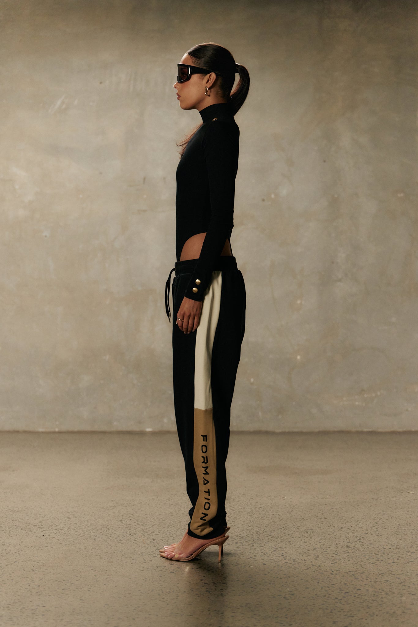 Side view of model wearing formation noir xaria bodysuit with gold-toned signature accents and noir xion track pants with drawstring and ivory and desert contrast panel with logo