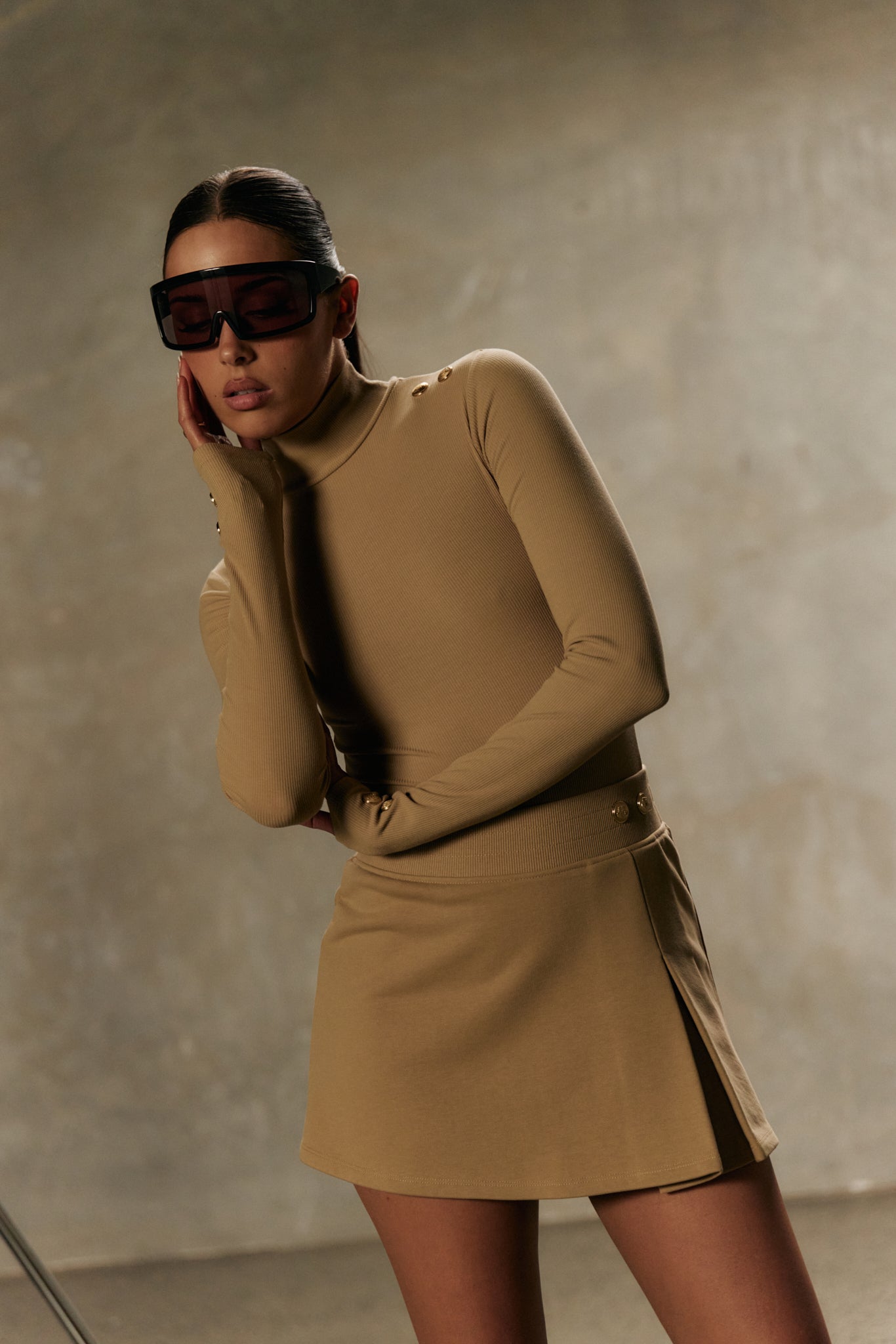 Model wearing formation desert xaria bodysuit with gold-toned signature accents and desert xya skirt with soft pleat and gold-toned signature accents