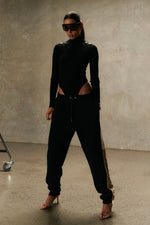 Model wearing Formation Xion noir track pants with straight leg, desert and ivory side panels, gold-toned accents and embroidery logo.