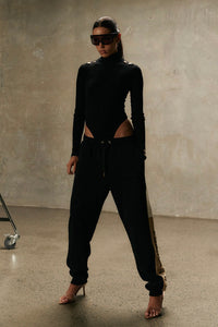 Model wearing Formation Xion noir track pants with straight leg, desert and ivory side panels, gold-toned accents and embroidery logo.